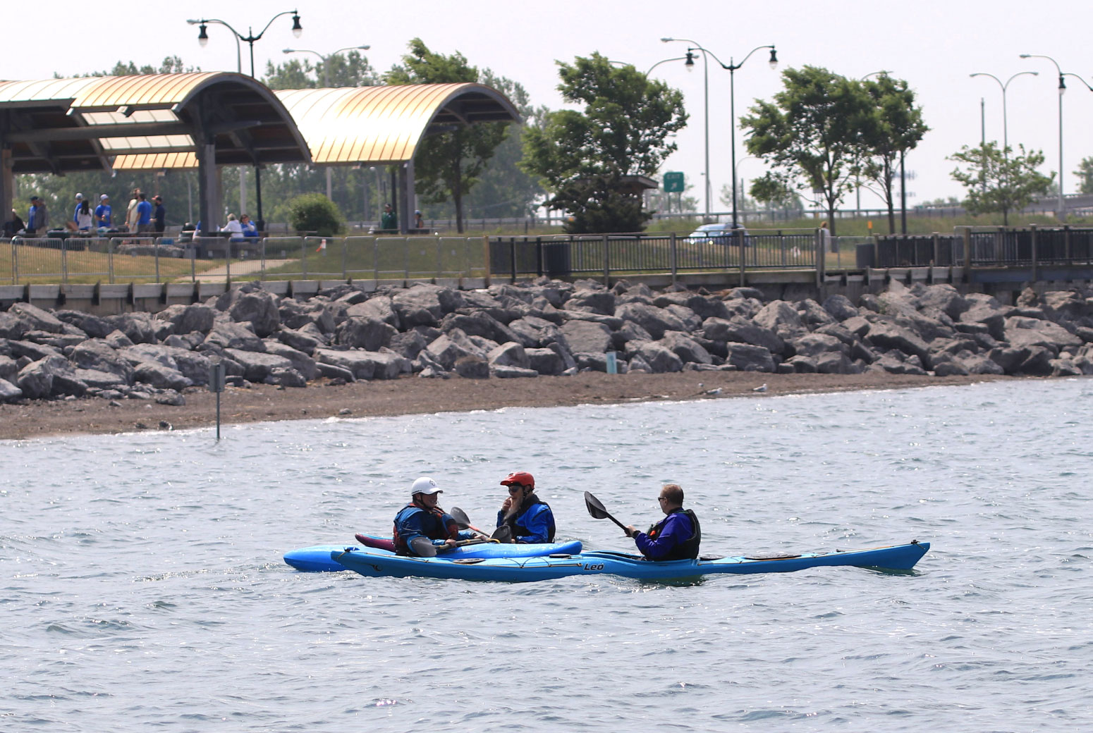 Three kayakers in the water in front of an outdoor event pavilion.