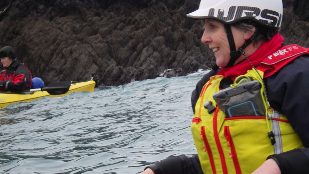 Close up of woman kayaking in rough water with rocky coast behind her